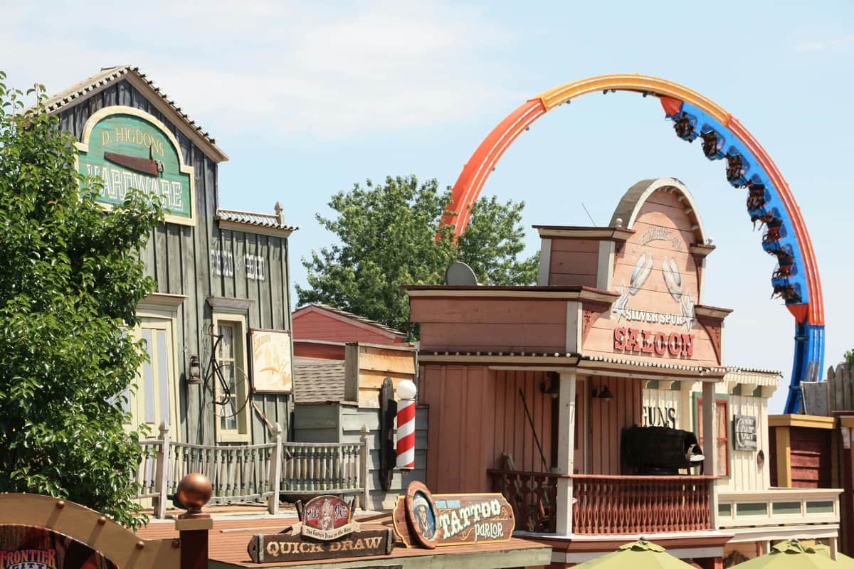 Six Flags Frontier City: Ein-Tages-Ticket Ein Vergnügungsparkbereich im Western-Stil mit Gebäuden im Saloon-Stil und einer farbenfrohen Achterbahn im Hintergrund.