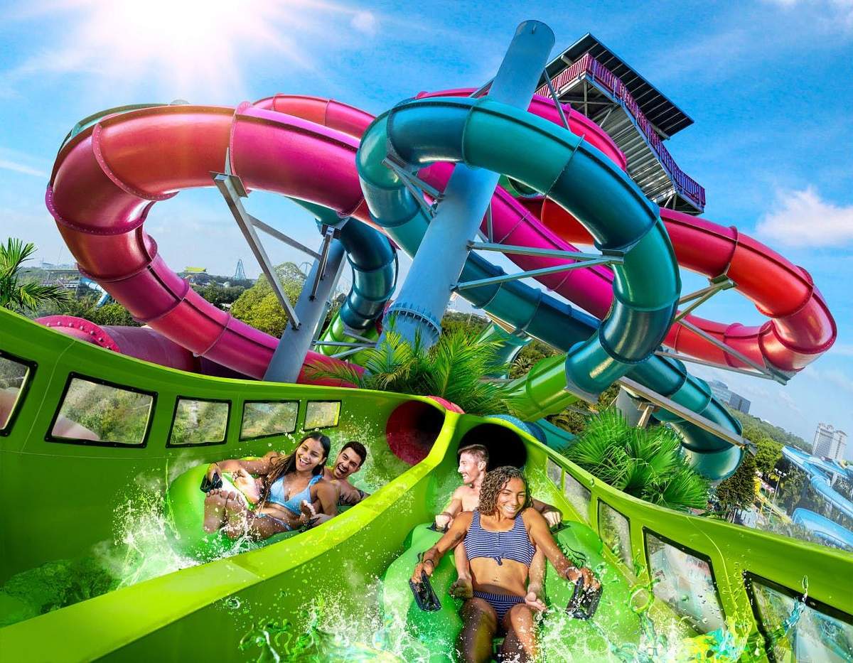 Four people descending a green waterslide with intertwined red, blue, and purple slides in the background under a clear blue sky.