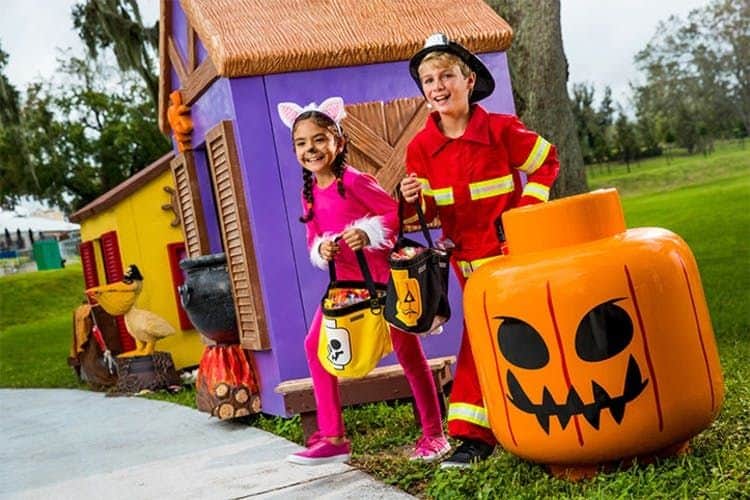 Two children in Halloween costumes, a pink cat and a firefighter, holding candy buckets in front of a playful, spooky house.