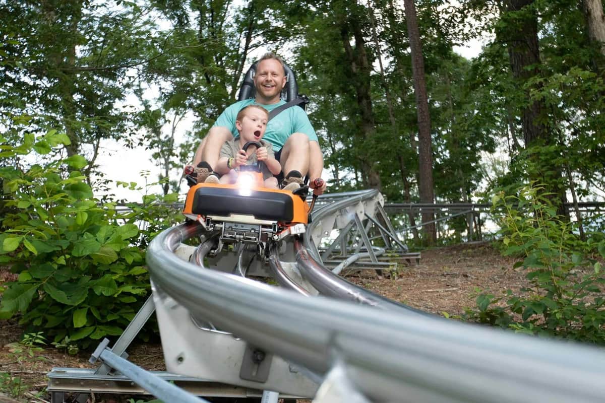 A man and child ride together on a roller coaster through a wooded area, both appearing excited and smiling.