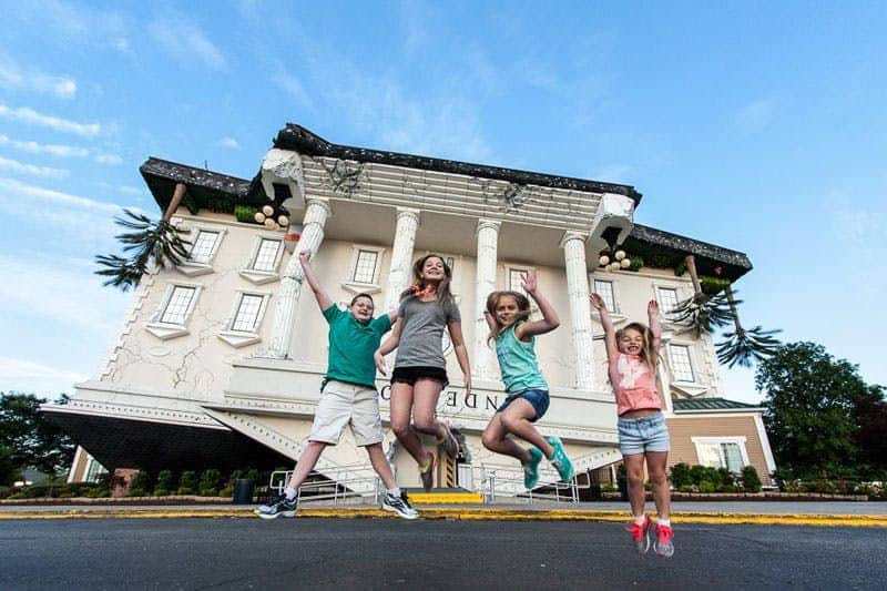 Four children jump in front of an upside-down building with columns on a sunny day.
