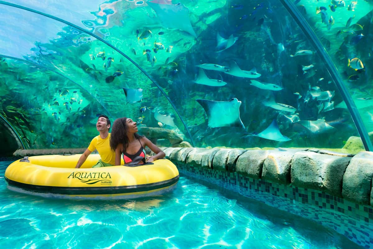 Two people in a yellow inflatable raft in an aquarium tunnel, surrounded by swimming stingrays and fish.