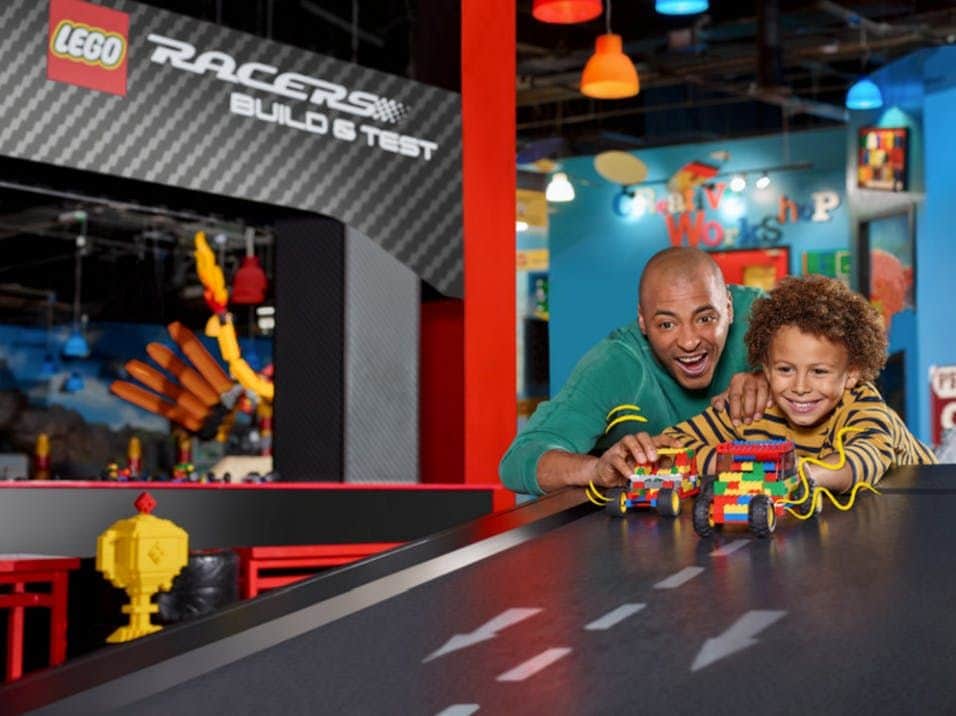 A man and a boy happily race colorful LEGO cars on a track in a playful, brightly-lit indoor environment.