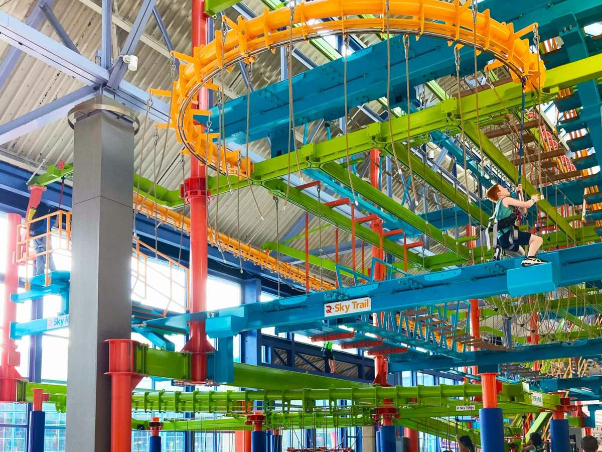 Indoor ropes course with colorful obstacles, safety harnesses, and ropes. A "Sky Trail" sign is visible, and one person is on the course.