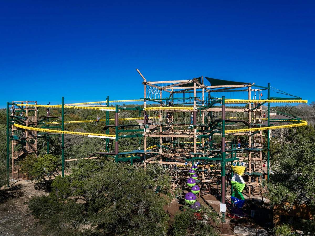 Twisted Trails at Natural Bridge Caverns: Entry Ticket Large outdoor ropes course with multiple levels, obstacles, and climbers. Trees and a clear blue sky in the background.
