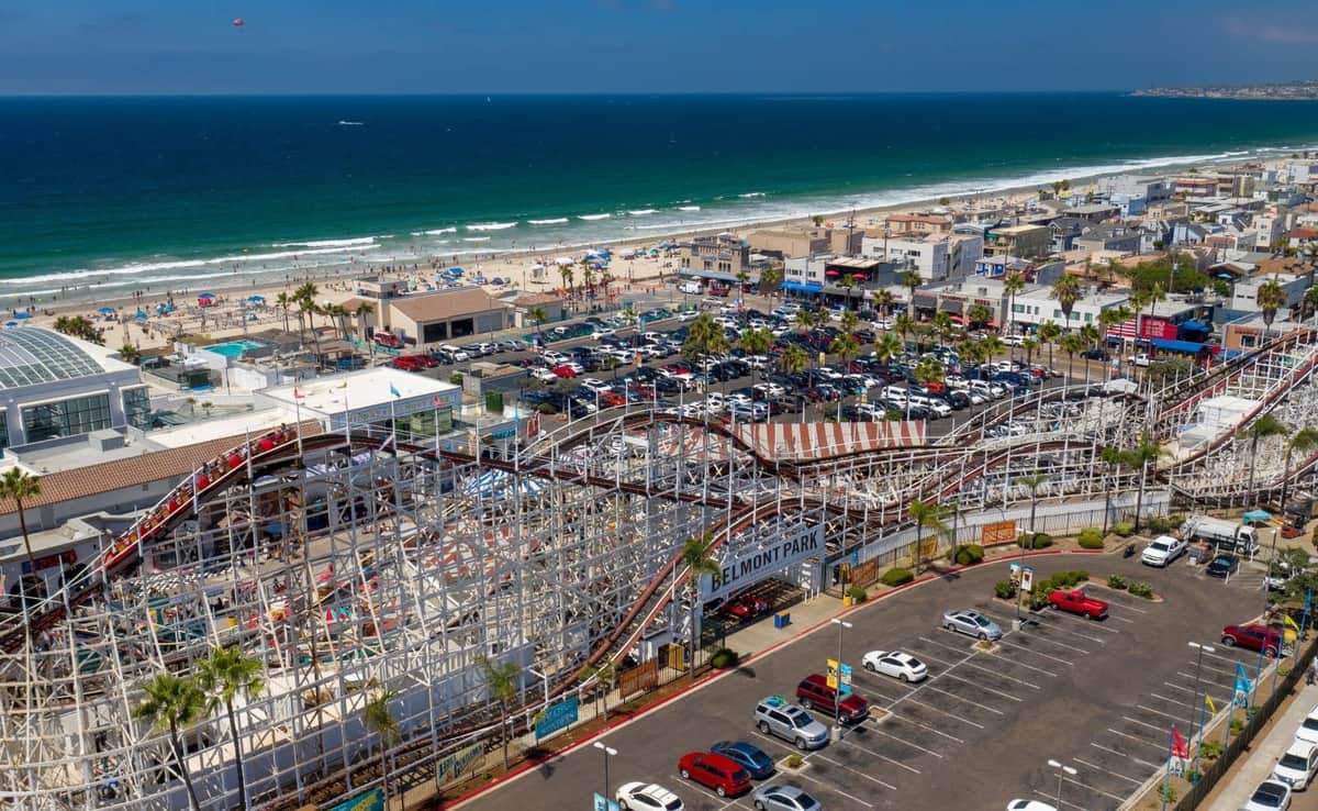 An aerial view of Belmont Park with a wooden roller coaster, parking lot, beachgoers, and the ocean in the background.
