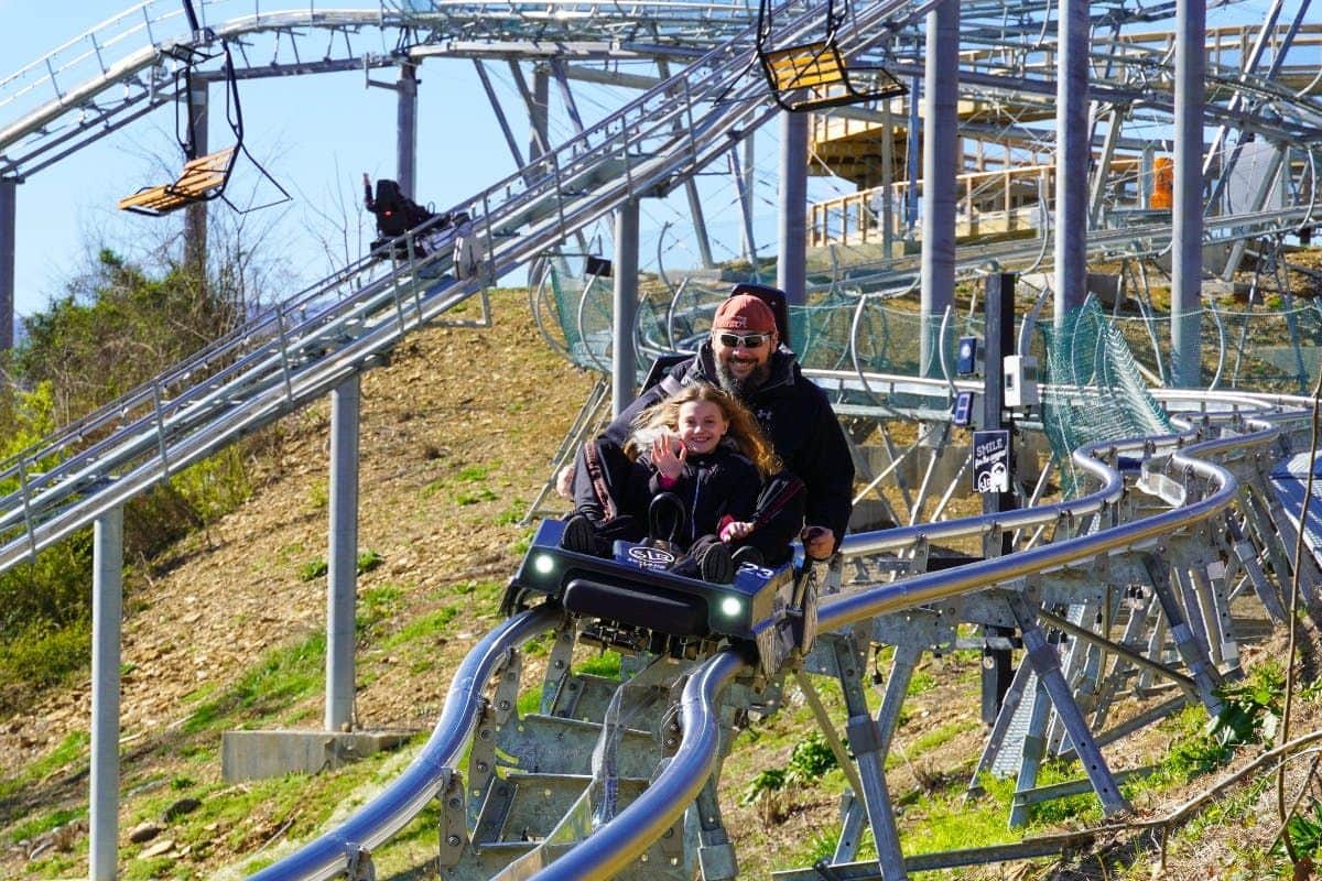 Two people smiling and waving while riding a personal roller coaster on a metal track with structure in the background.