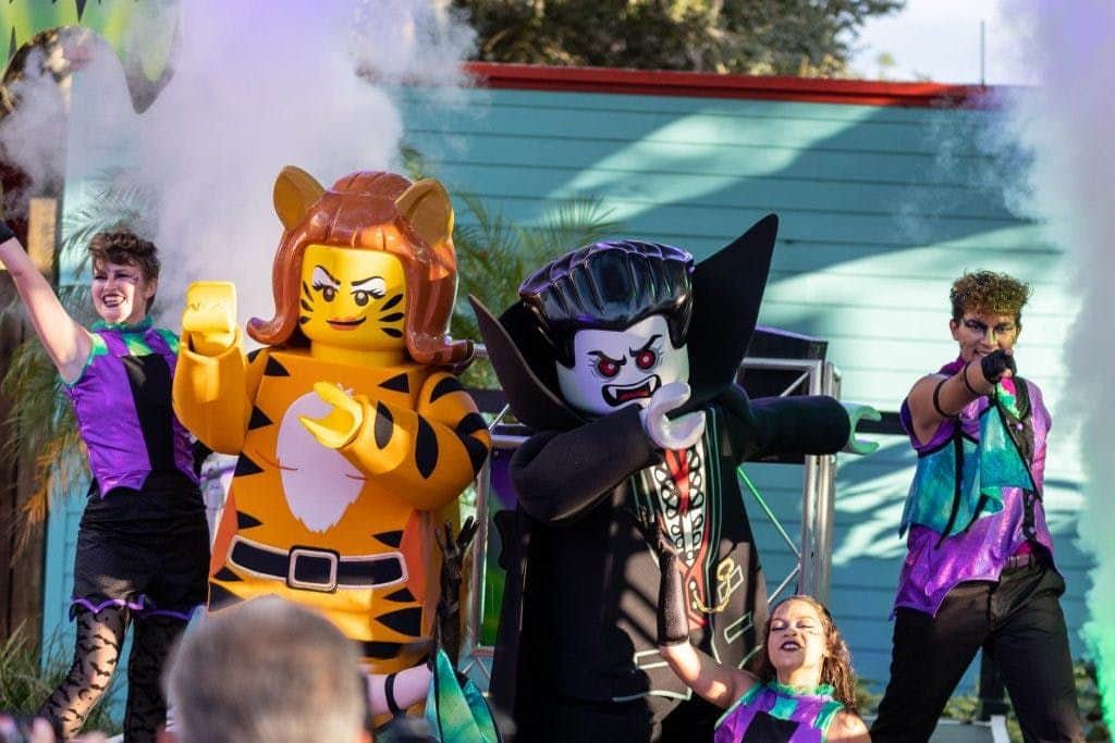 Large LEGO figures of a tiger and a vampire, surrounded by costumed people and stage lights, with smoke in the background.