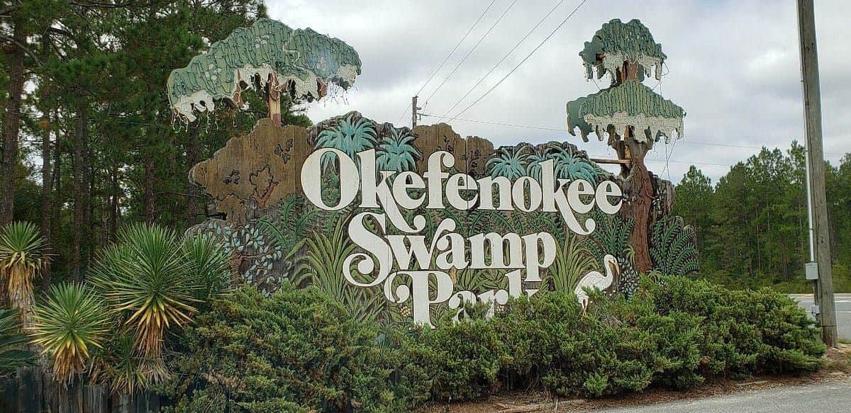 Sign that reads "Okefenokee Swamp Park" surrounded by illustrated trees and plants, with a bird illustration on the right.