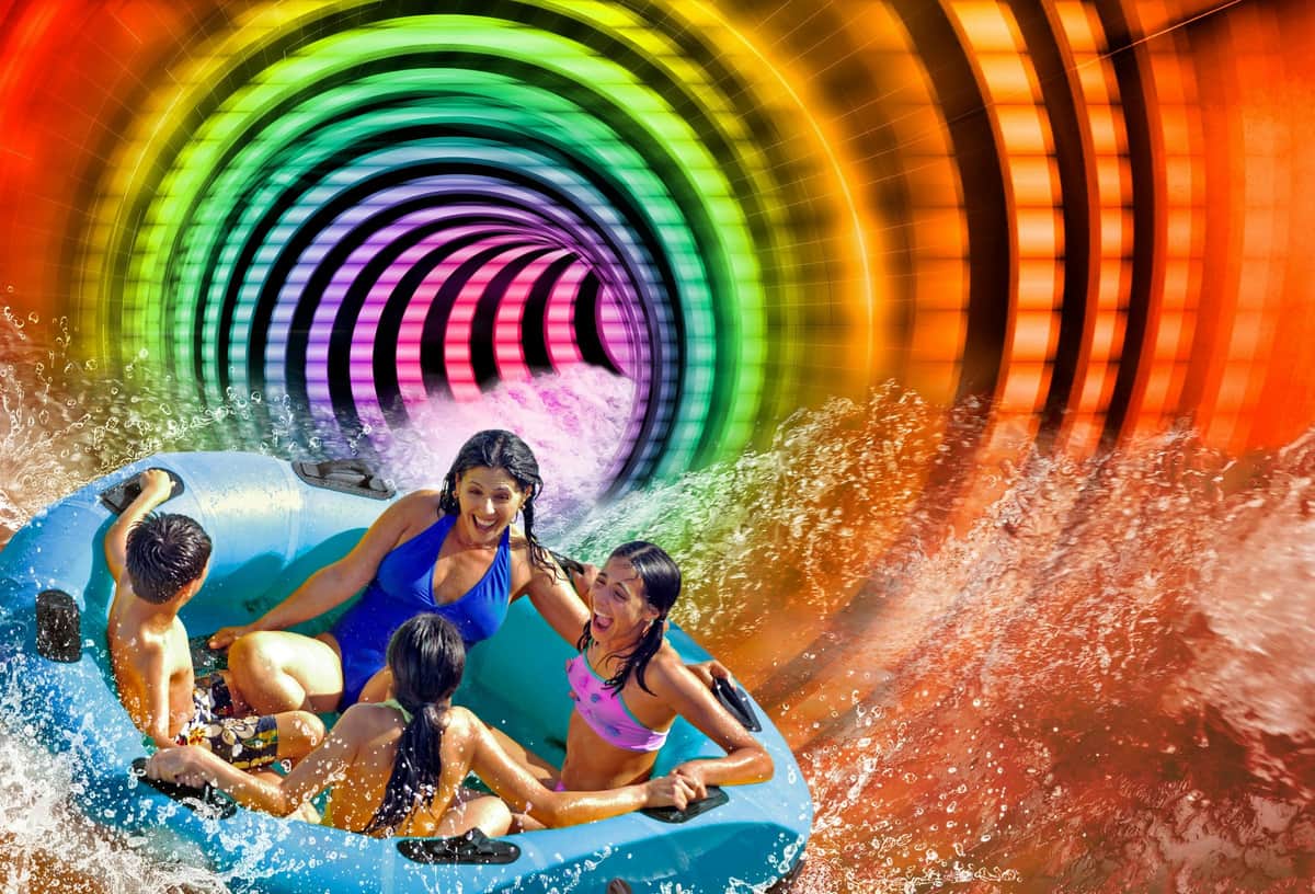 Four people in swimsuits ride a blue inflatable raft through a colorful, spiraling water slide, splashing water around them.