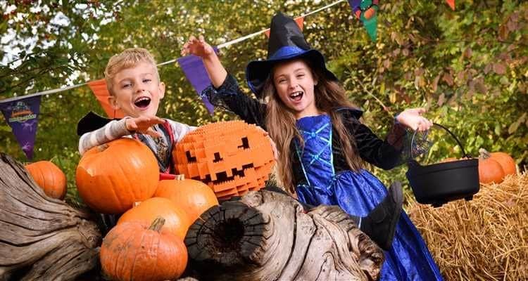 A child smiles surrounded by pumpkins and a LEGO jack-o’-lantern, with festive flags and trees in the background.