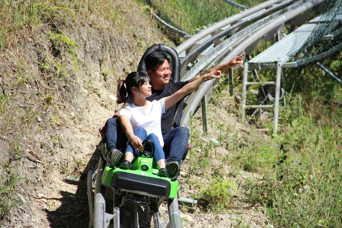 Two people riding a green sled on a mountain coaster track surrounded by grass and vegetation, both pointing ahead.