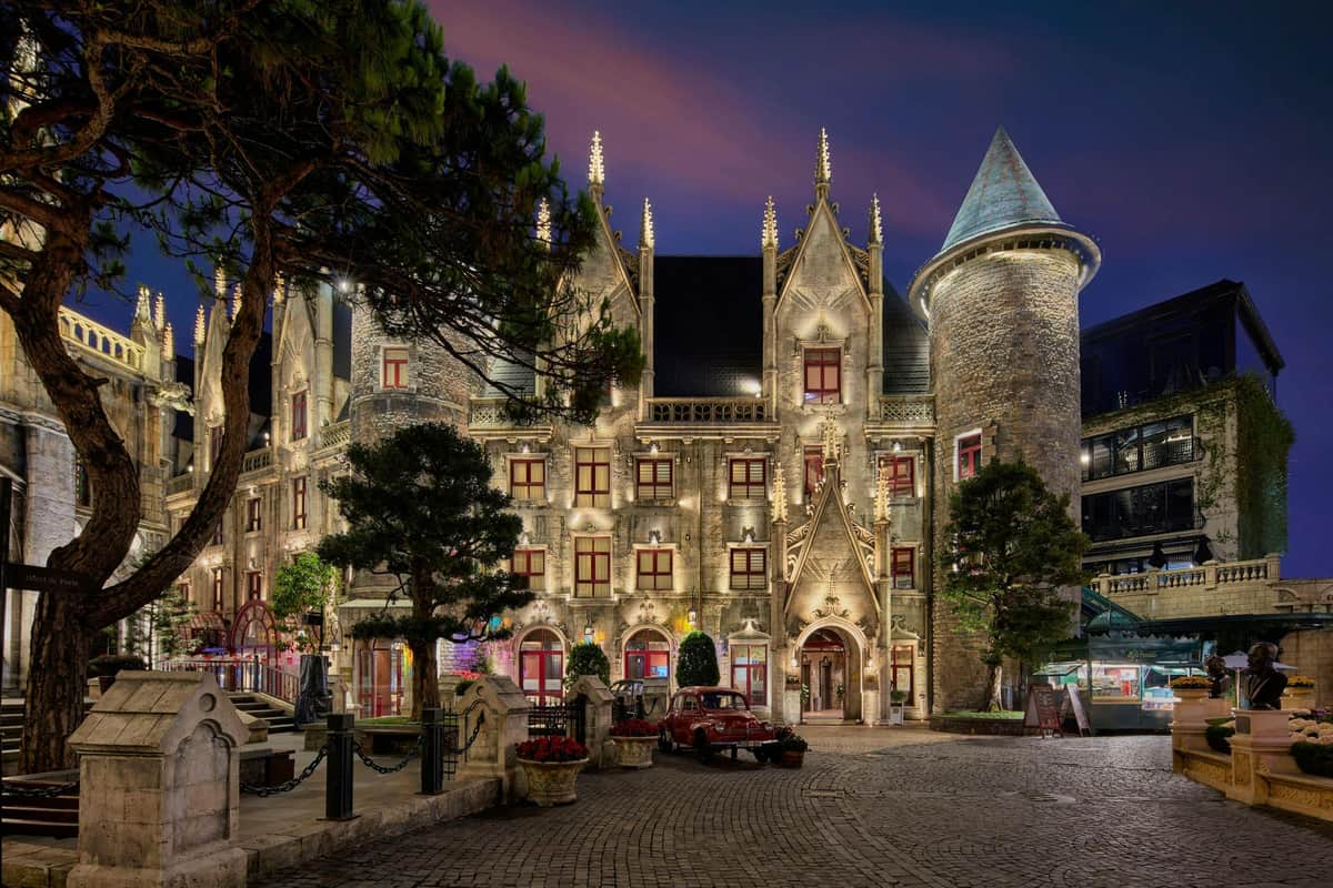 Illuminated castle-like building with pointed towers, arched entrance, and courtyard at night, framed by trees.