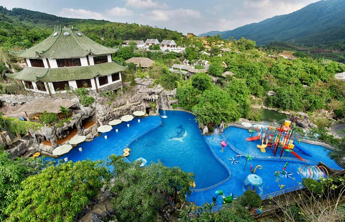 A scenic view of a large outdoor pool surrounded by greenery, a traditional-style building, and a colorful play area.