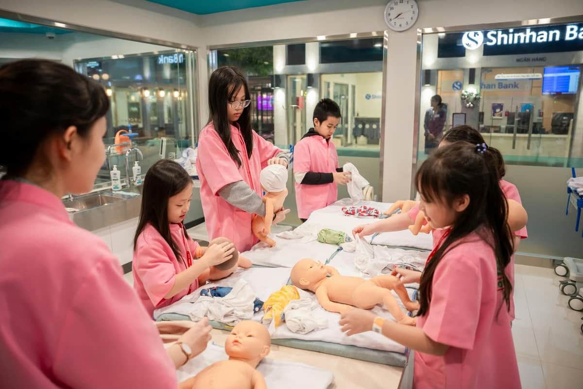 Children wearing pink uniforms practicing caregiving skills with baby dolls in a brightly lit classroom.