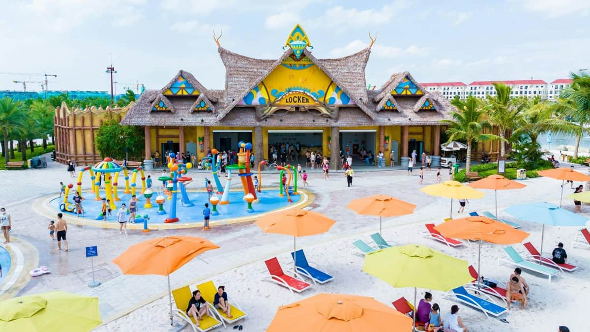 A vibrant tropical beach scene with palm trees, colorful umbrellas, and people enjoying a lagoon and sandy shore.