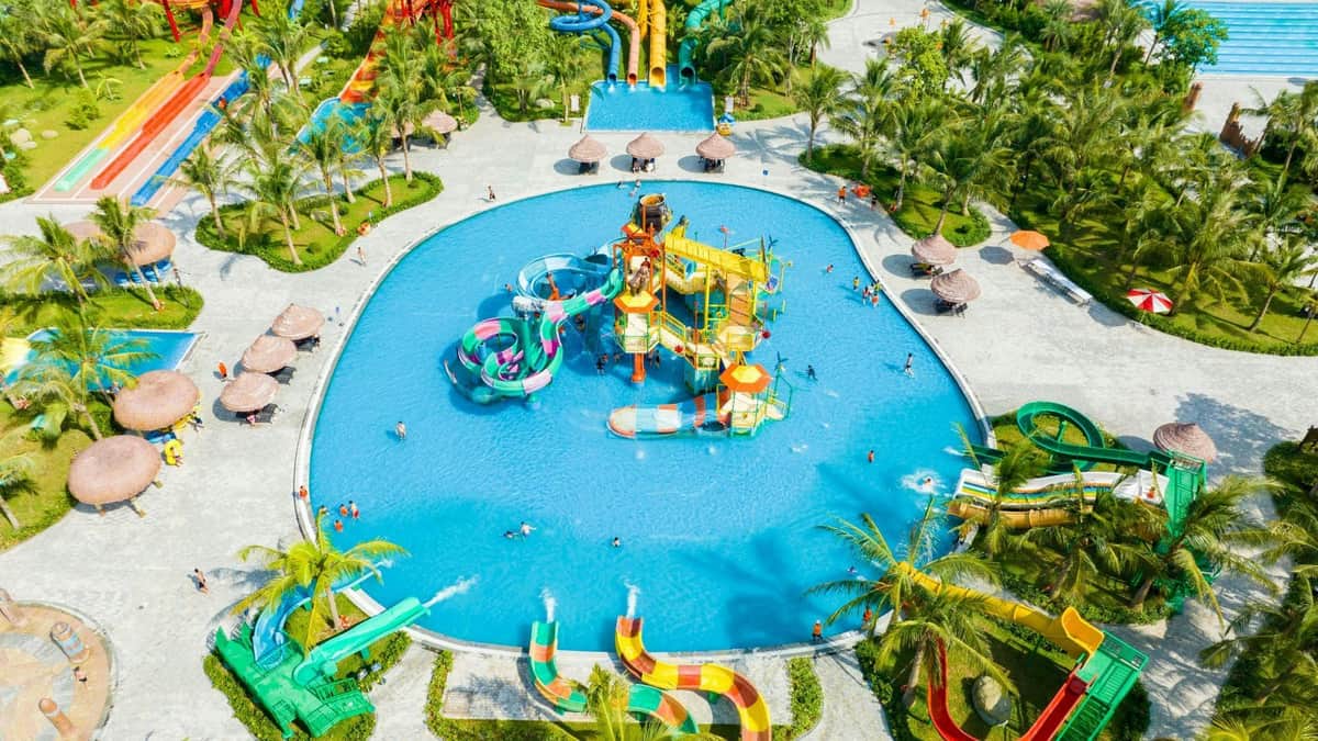 A colorful water park with slides, a pool, and palm trees. People are enjoying various activities under a bright sky.