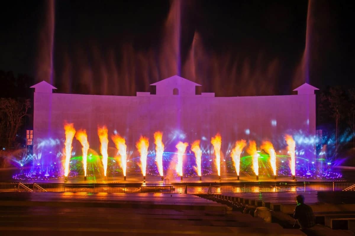 A nighttime water and light show with colorful fountains, illuminated sprays, and fire effects in front of a building.