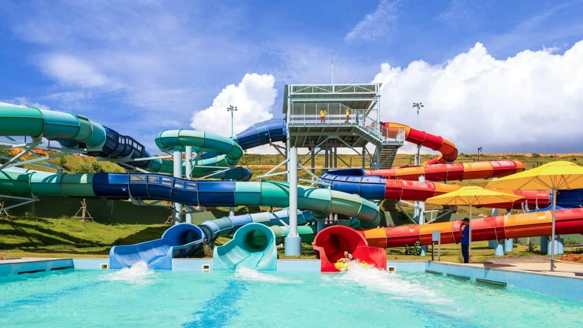 Colorful water slides leading into a pool under a blue sky, with a platform and people visible at the slide entrances.
