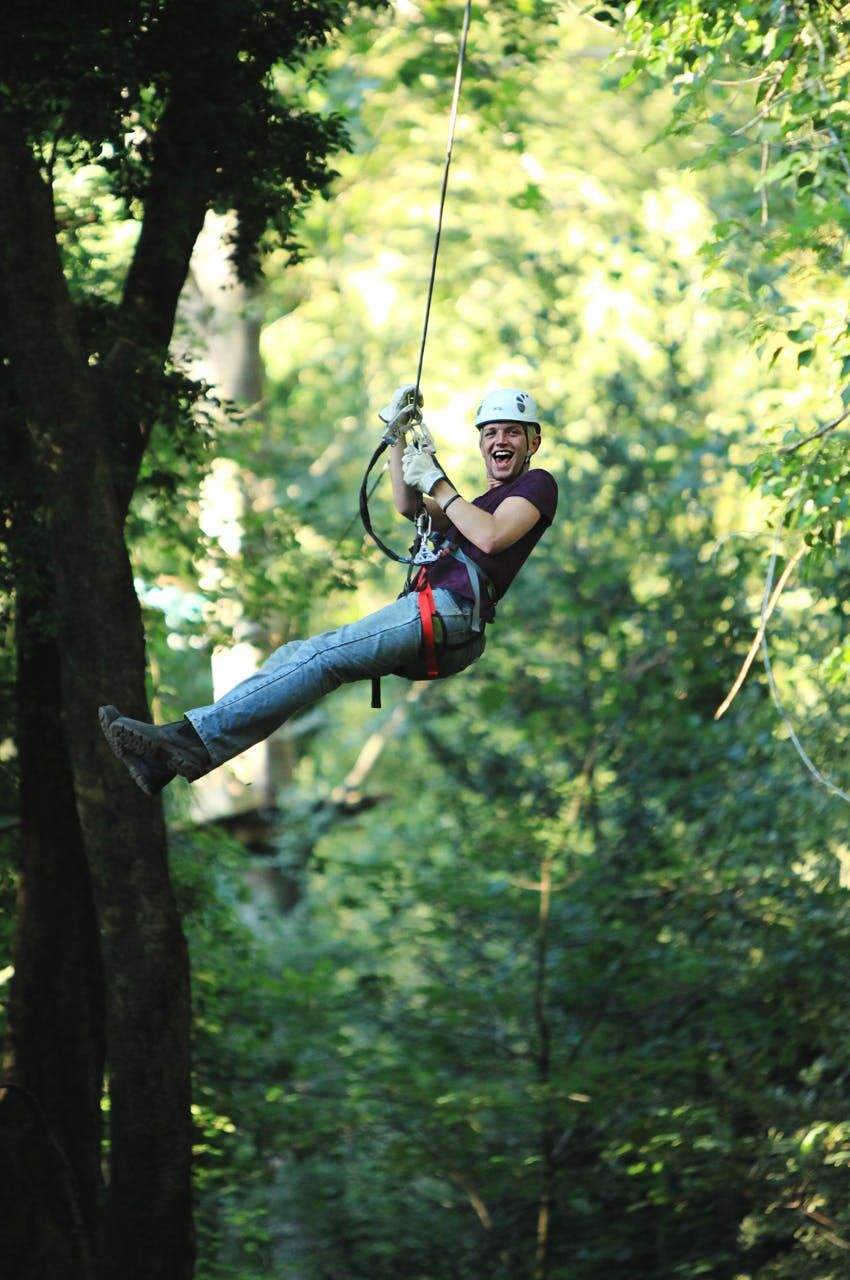 Person ziplining through a forest, wearing a helmet and a harness, smiling with an excited expression. Trees and greenery in the background.
