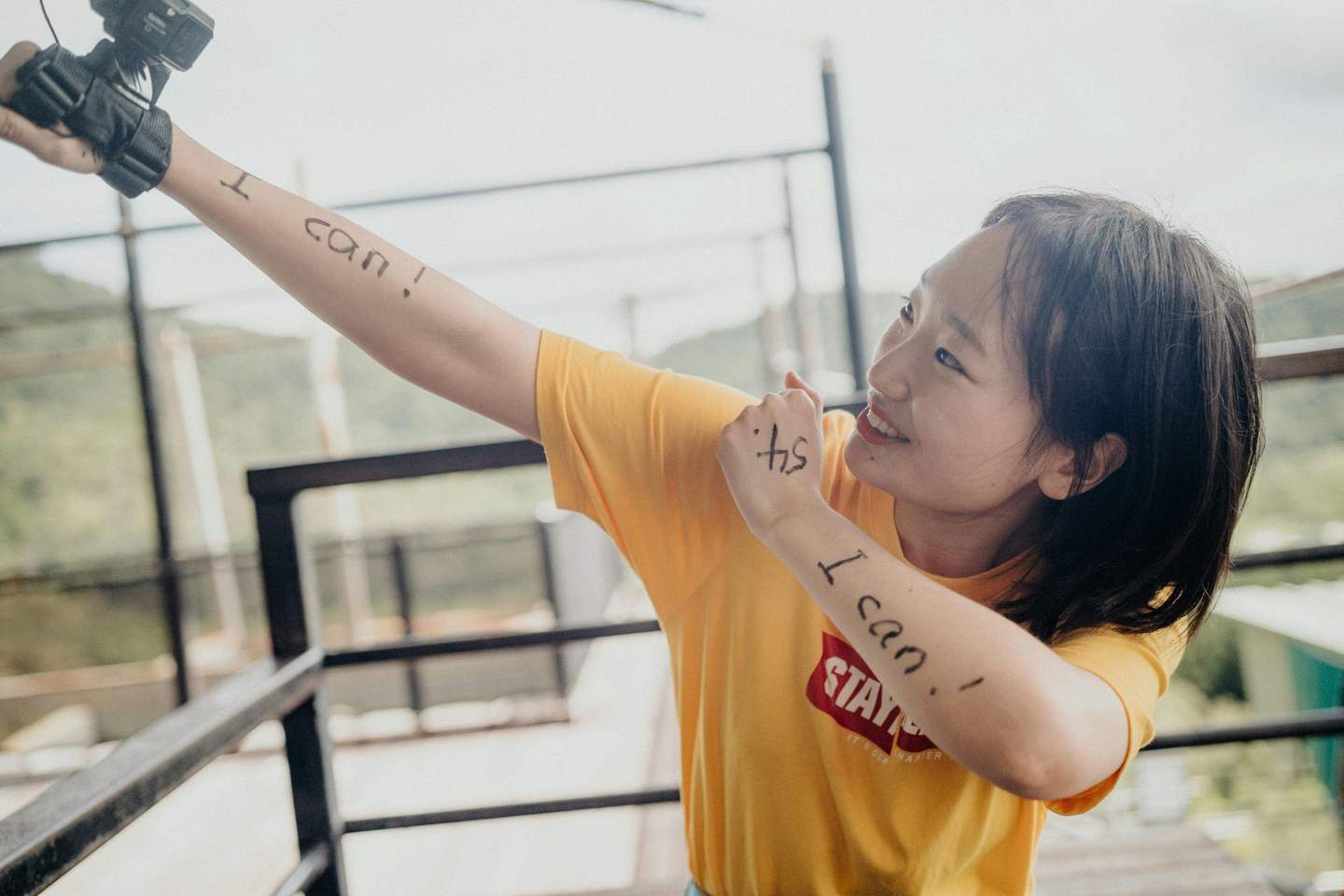 Skypark Cairns por AJ Hackett Una persona con una camiseta amarilla que lleva escrito "¡Yo puedo!" en los brazos sostiene una cámara, sonriendo enérgicamente al aire libre.