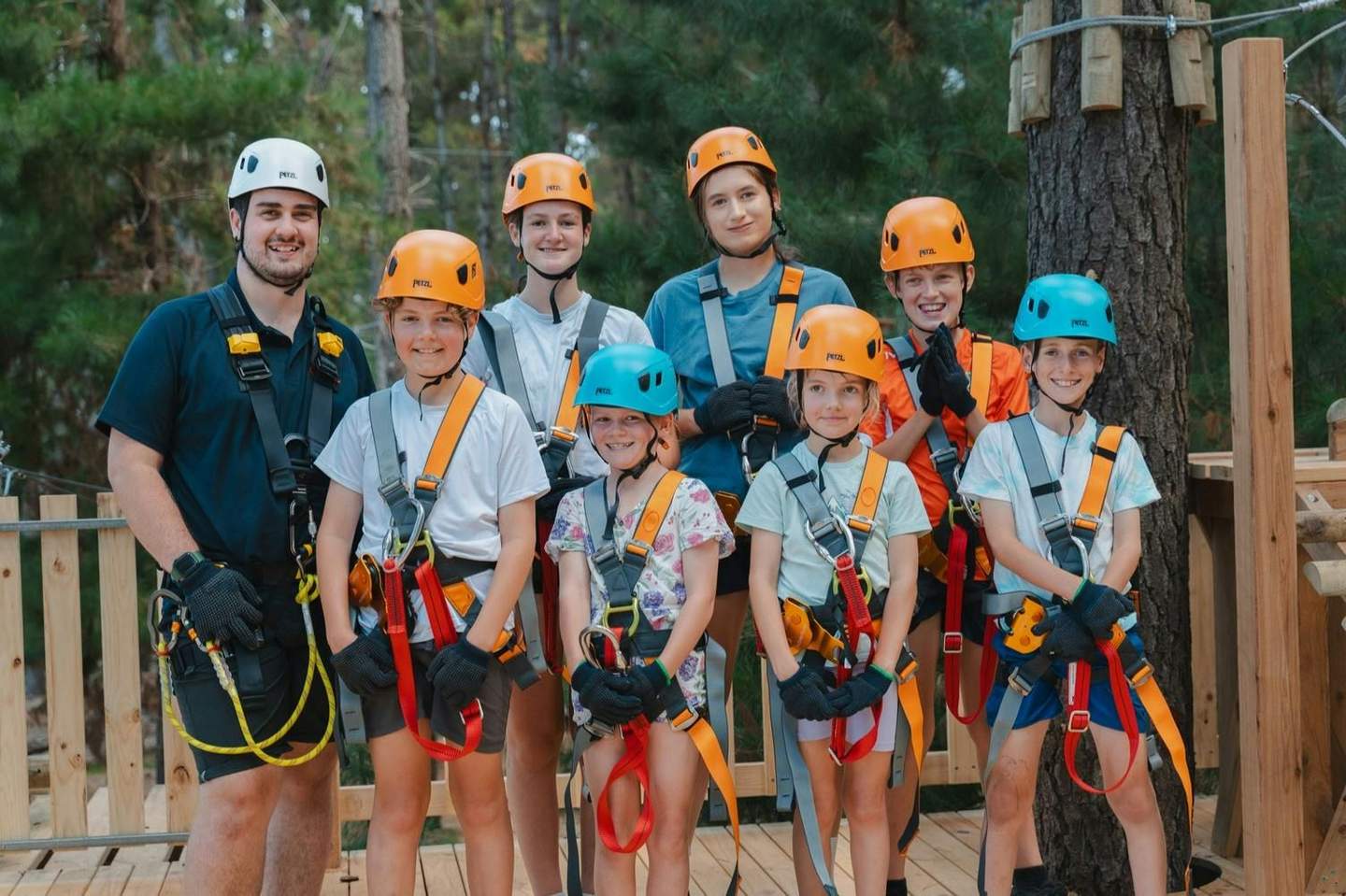 A group of people, including children and adults, wearing helmets and harnesses, standing outdoors on a wooden platform.