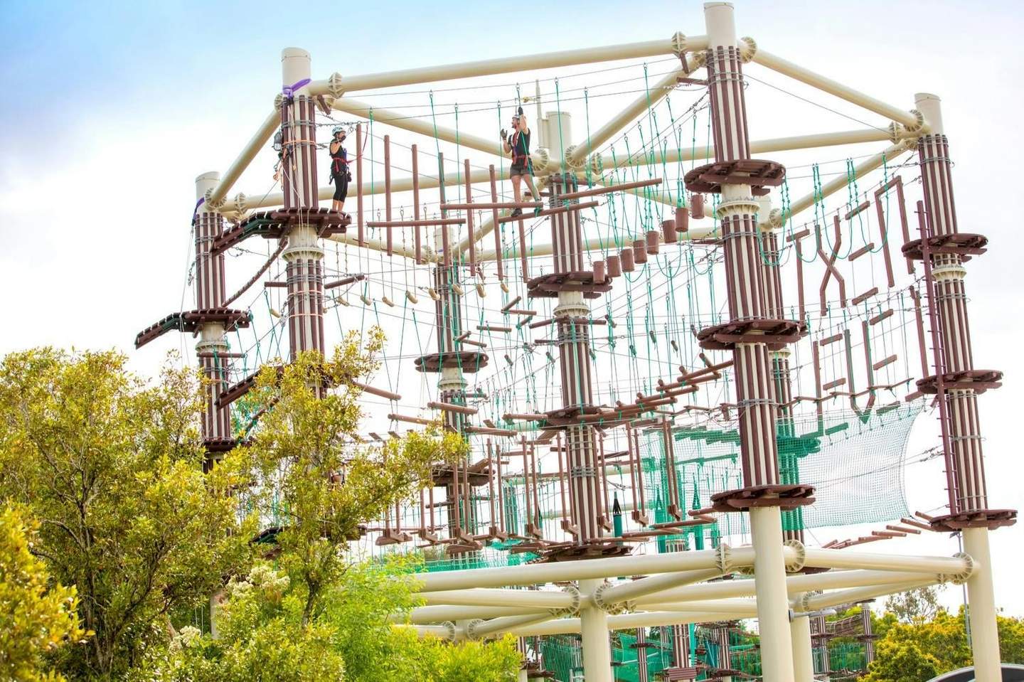 Two people on a high ropes course, navigating various suspended obstacles. The structure is surrounded by green foliage.