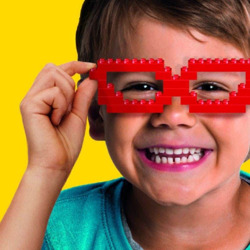 A child smiling while wearing red glasses made of LEGO bricks, against a bright yellow background.