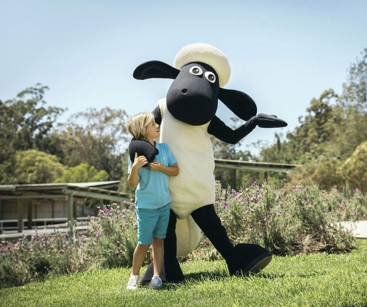 Paradise Country A smiling child in blue stands next to a large sheep mascot on a grassy area with flowers, trees, and clear sky in the background.