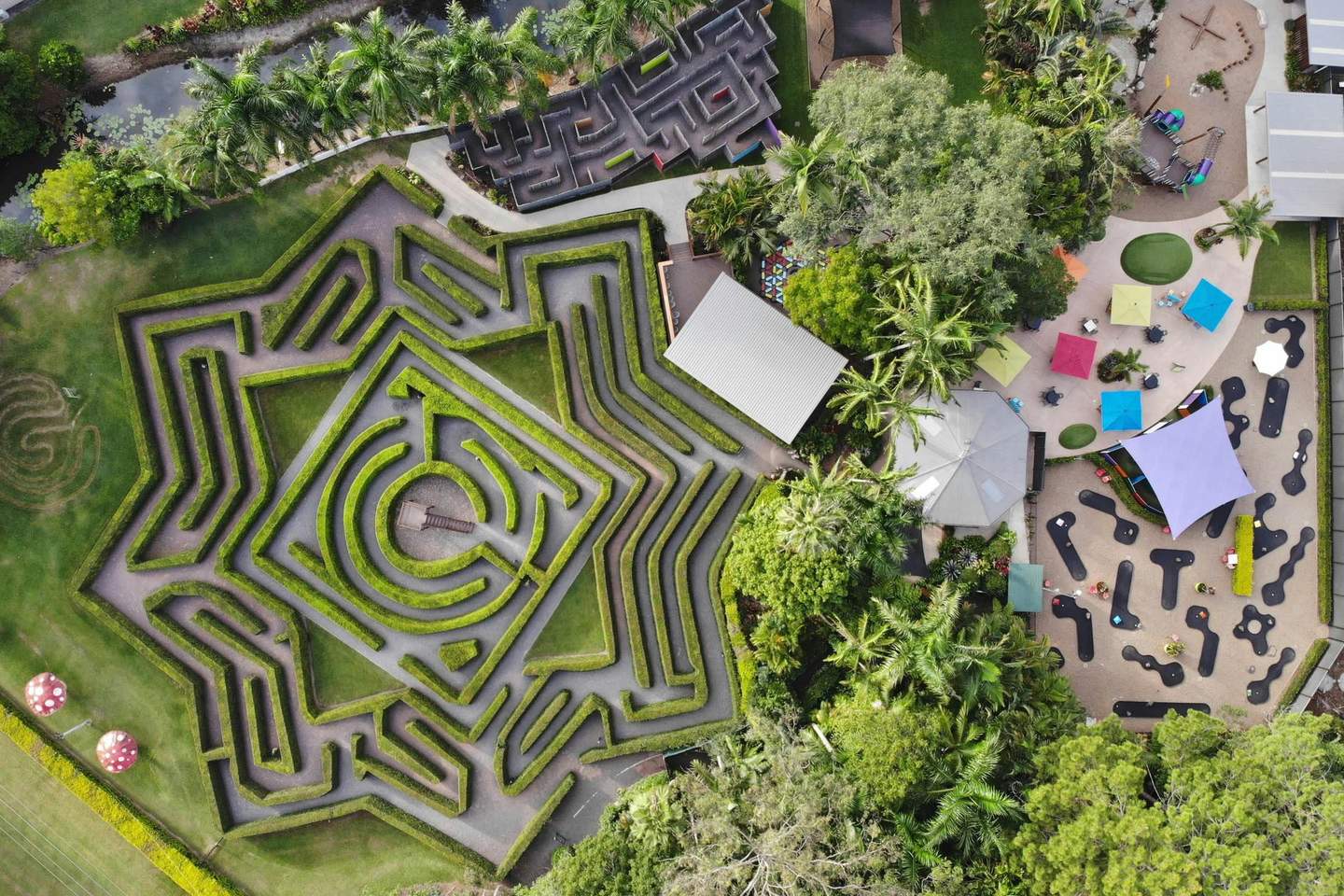 Aerial view of a geometric hedge maze, playground with colorful umbrellas, and surrounding trees and paths.