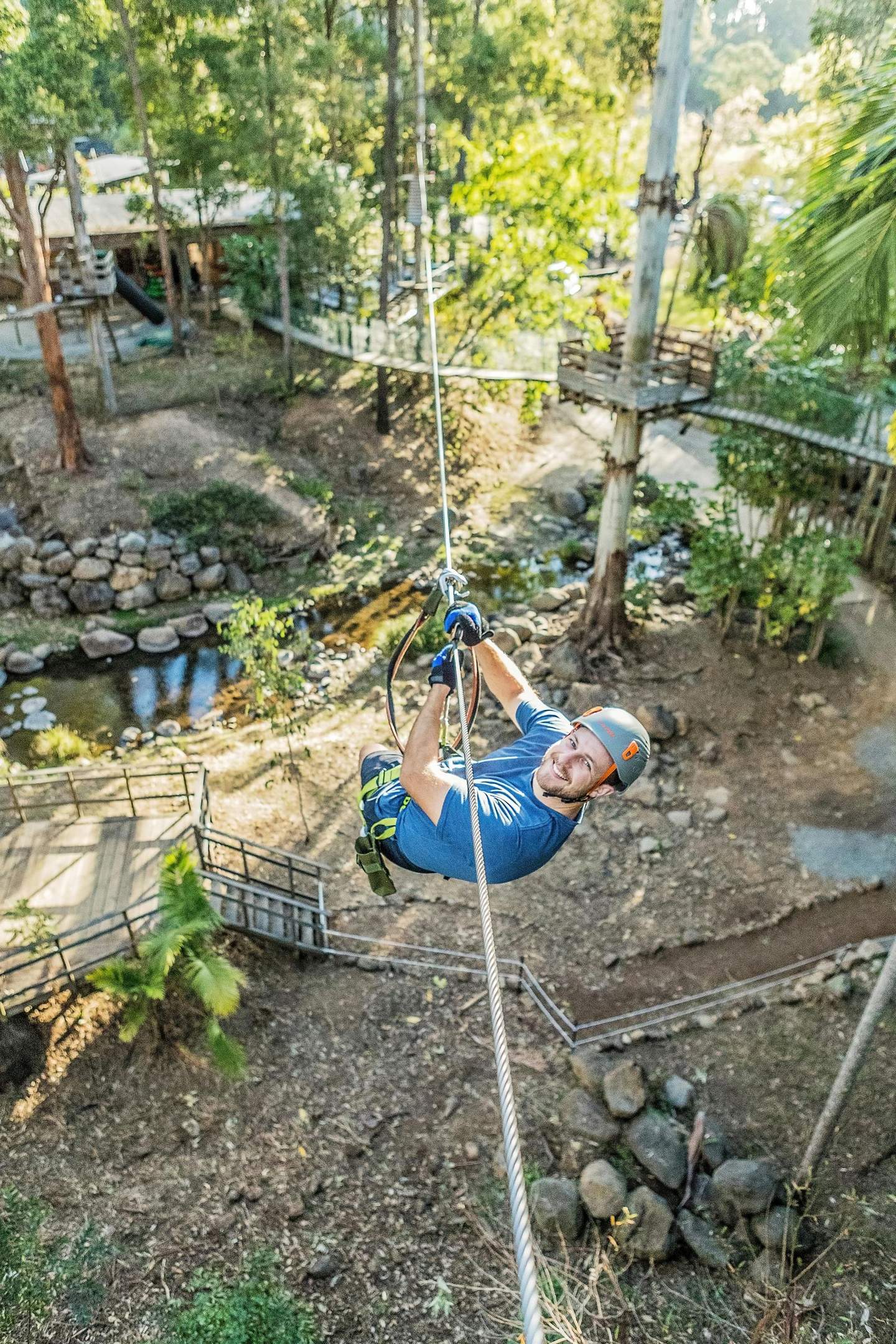 A man wearing a helmet and safety gear smiles while ziplining through a lush, wooded area with trees and a small stream below.