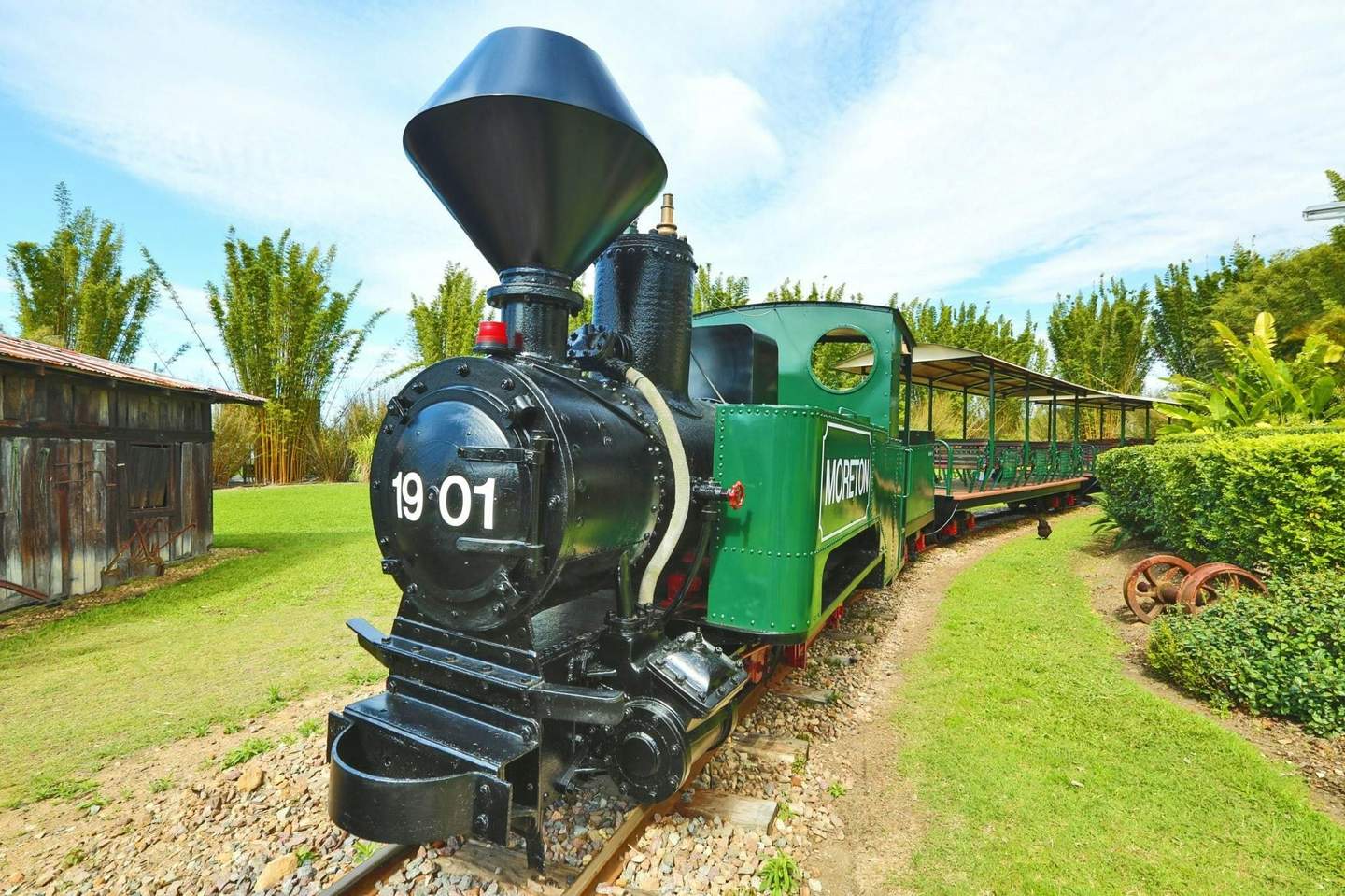 Vintage green steam locomotive with "1901" on the front and "MORETON" on the side, parked on tracks in a lush garden setting.