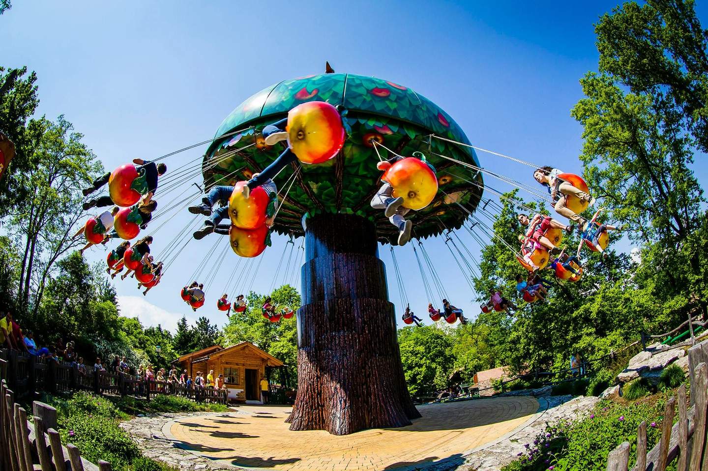 People riding a swing carousel with apple-shaped seats, set against a backdrop of lush trees and a clear blue sky.