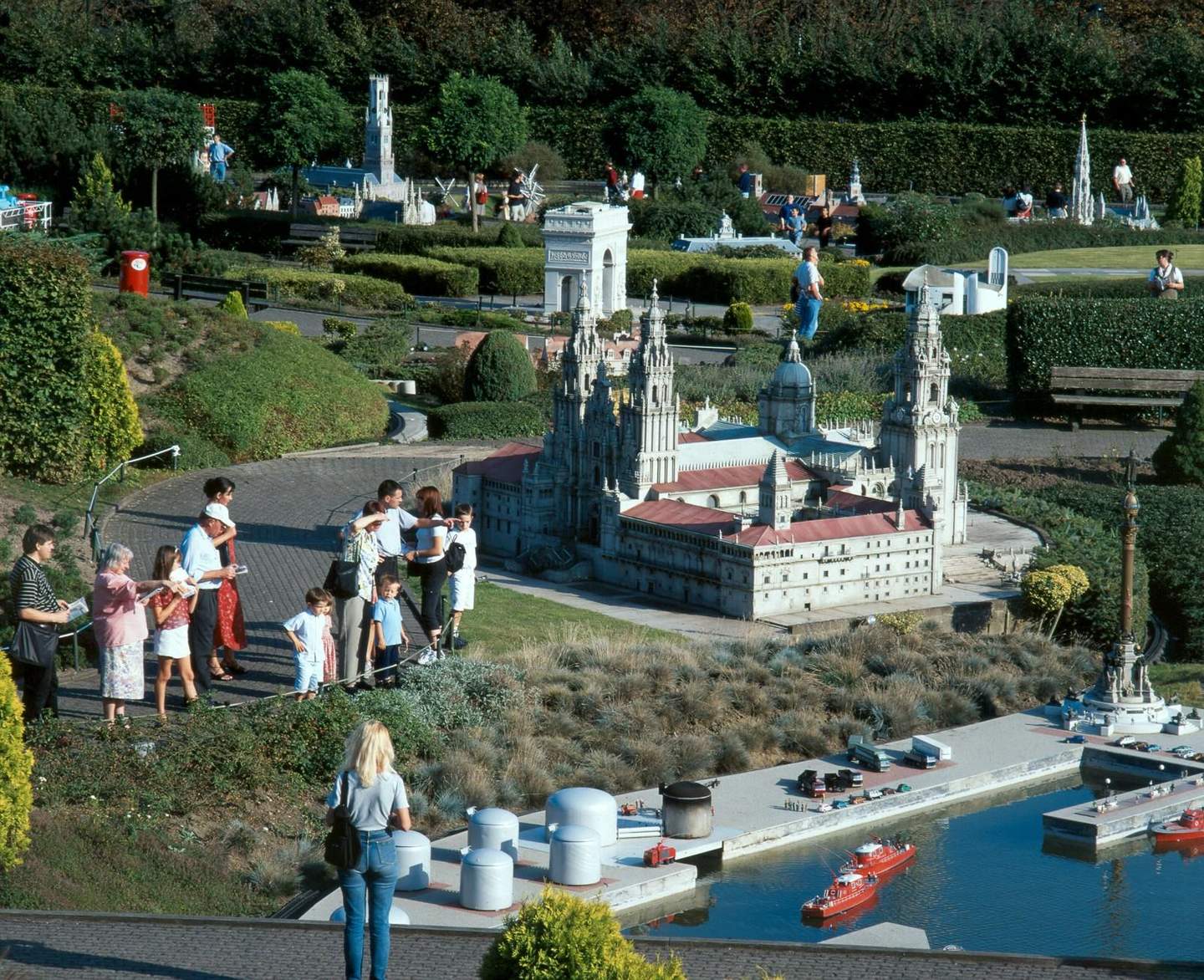 People admire detailed architectural miniatures in a lush park setting, featuring iconic landmarks and a small waterway with model boats.