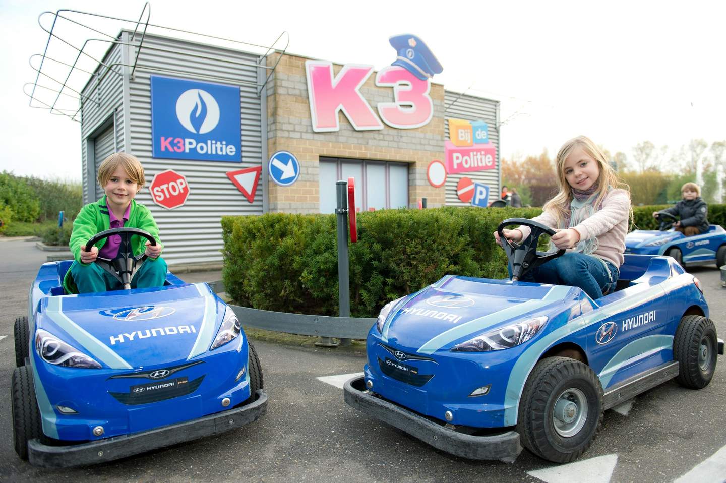 Two children driving blue Hyundai toy cars in front of a building with "K3 Politie" and traffic signs.