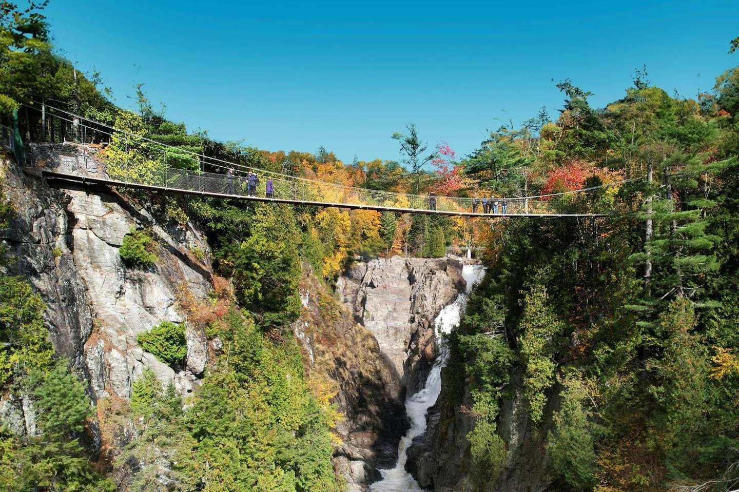 A suspension bridge spans above a gorge with a waterfall, surrounded by forested cliffs and autumn foliage.