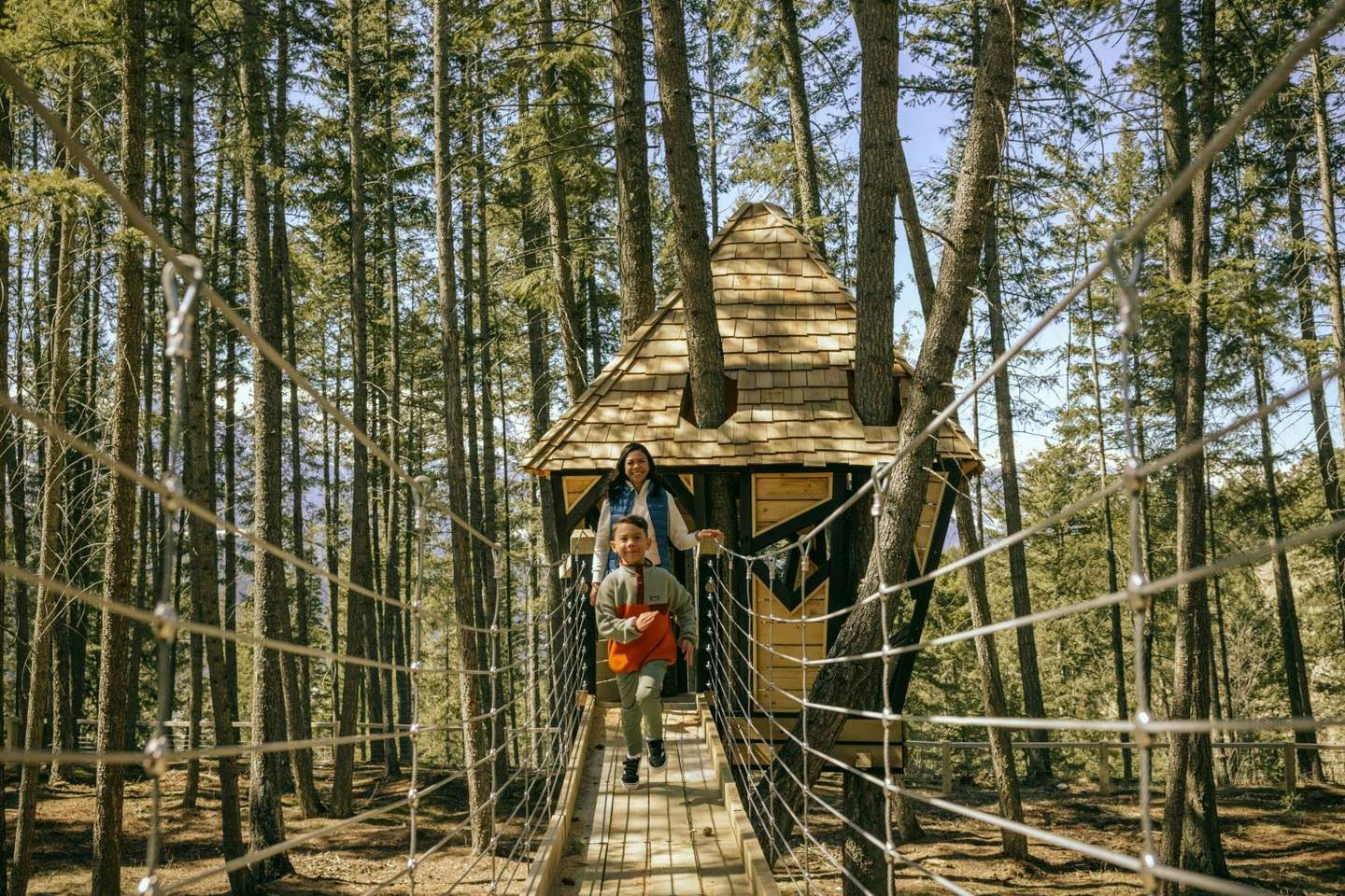 A child and an adult are on a rope bridge leading to a wooden treehouse in a forest. The weather is sunny.