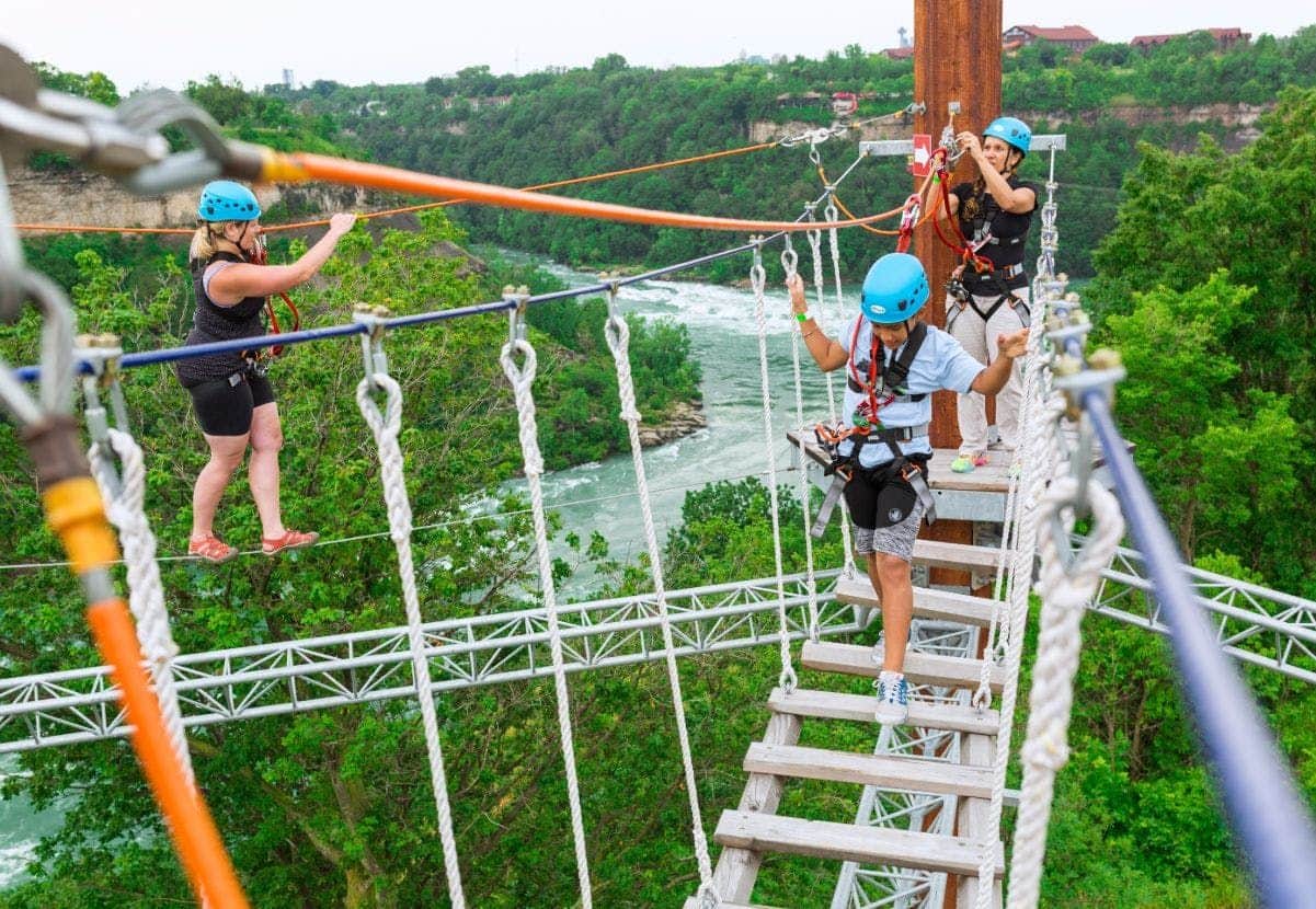 People wearing helmets and harnesses traverse a rope bridge with a river and forested landscape in the background.