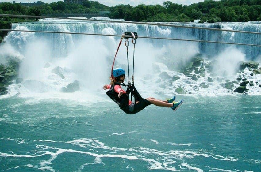 A person in a blue helmet and harness ziplining over a powerful waterfall with lush greenery in the background.
