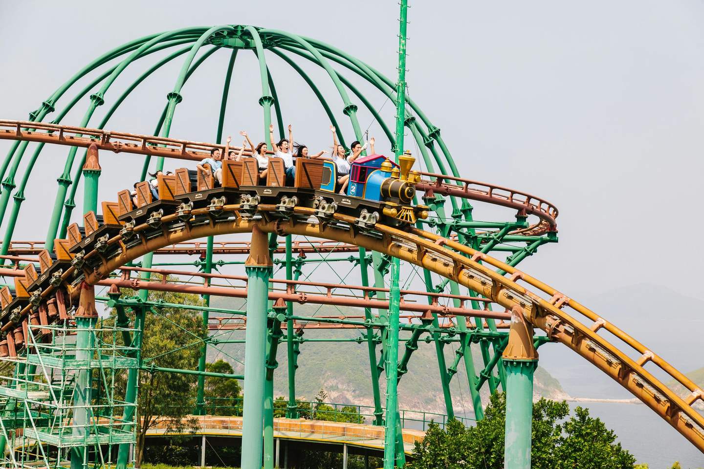 People riding a rollercoaster with green and brown tracks, set against a backdrop of trees, metal structures, and distant hills.