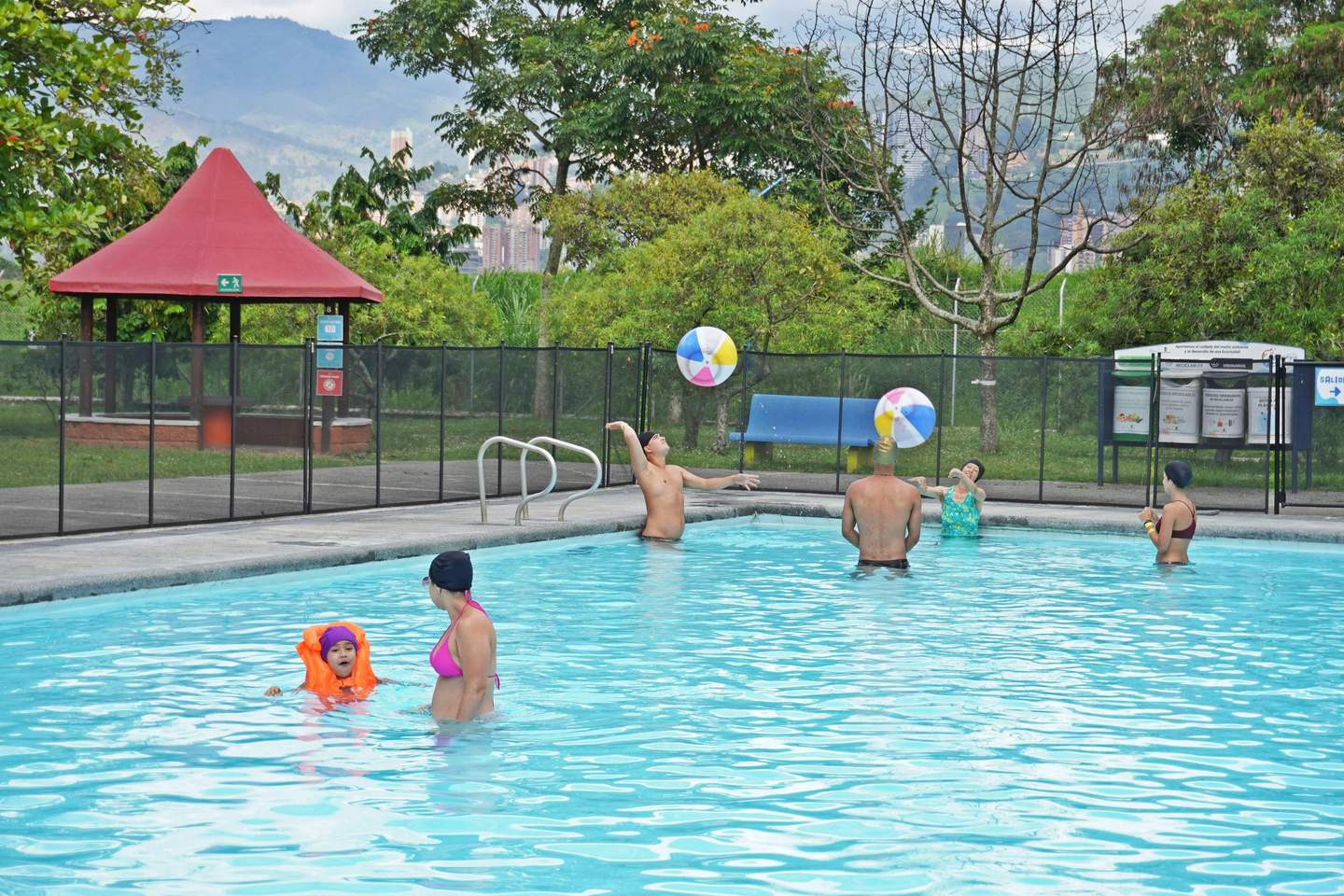 People playing with colorful beach balls in an outdoor swimming pool surrounded by trees, with a red-roofed gazebo in the background.