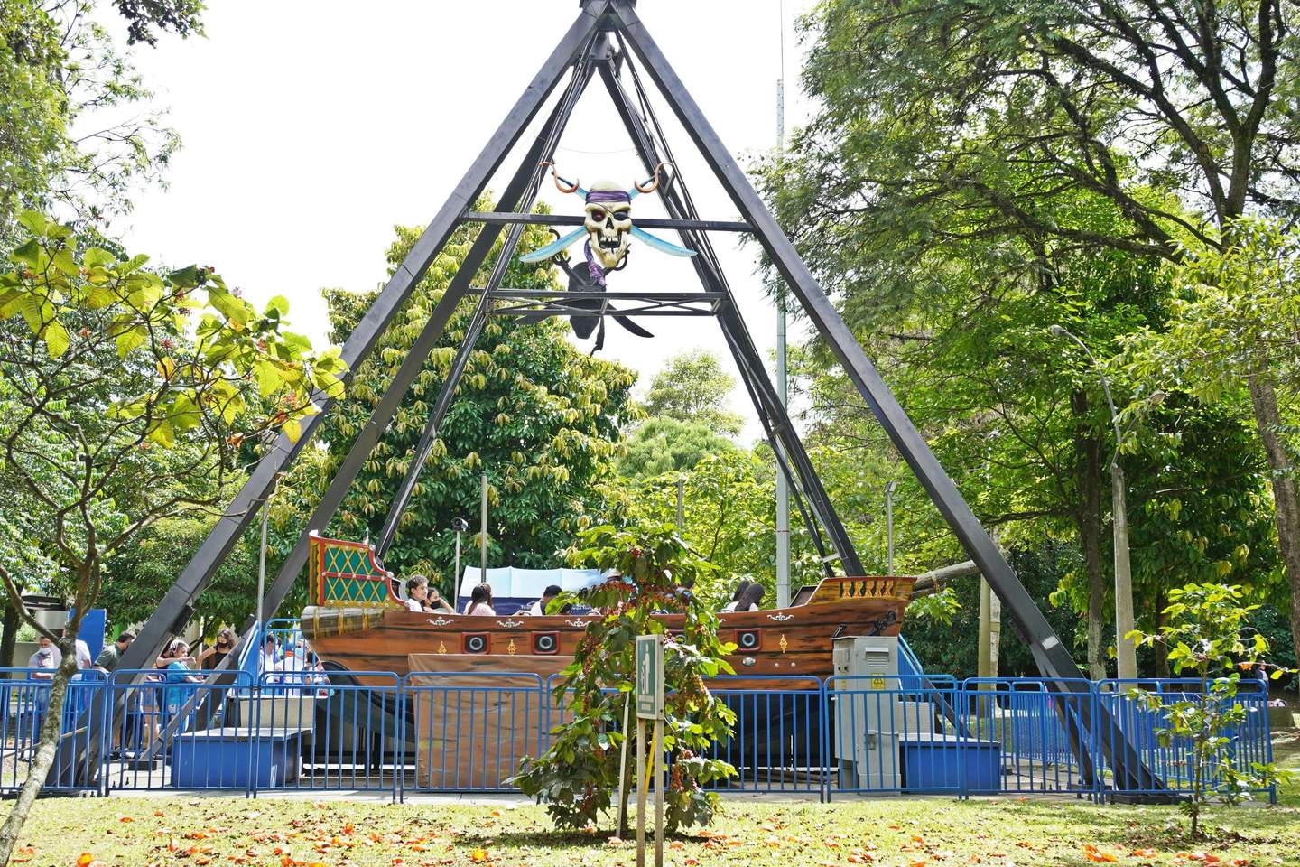 Pirate ship ride at an amusement park with passengers, surrounded by greenery and enclosed by blue metal railings.