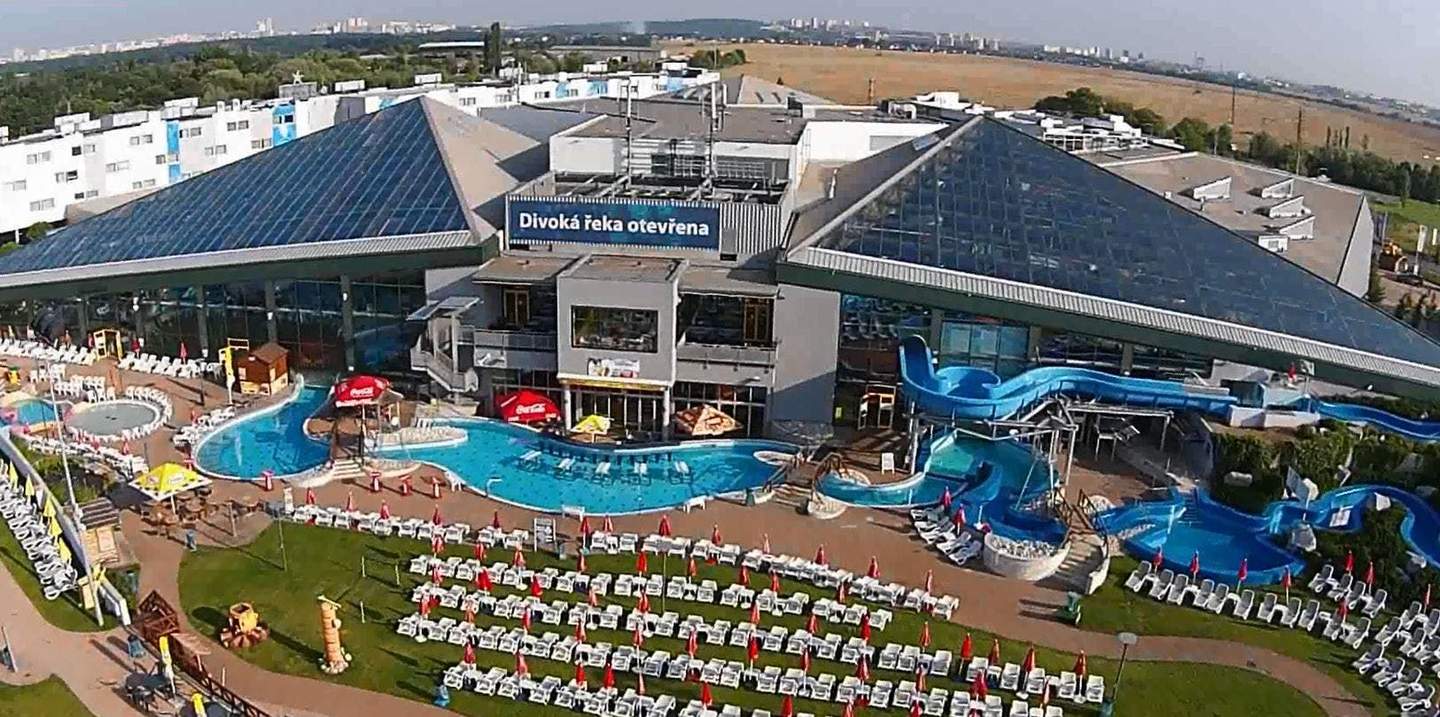 Large complex with pools, slides, and numerous sun loungers with umbrellas. Pyramid-shaped glass roof and building in background.