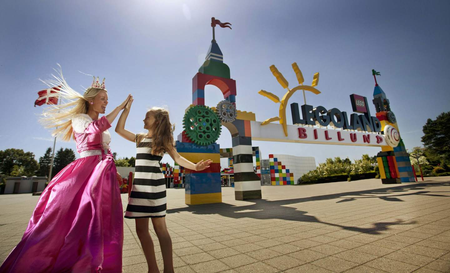 A woman in a pink dress and crown high-fives a girl in a black and white striped dress in front of the LEGOLAND Billund entrance.