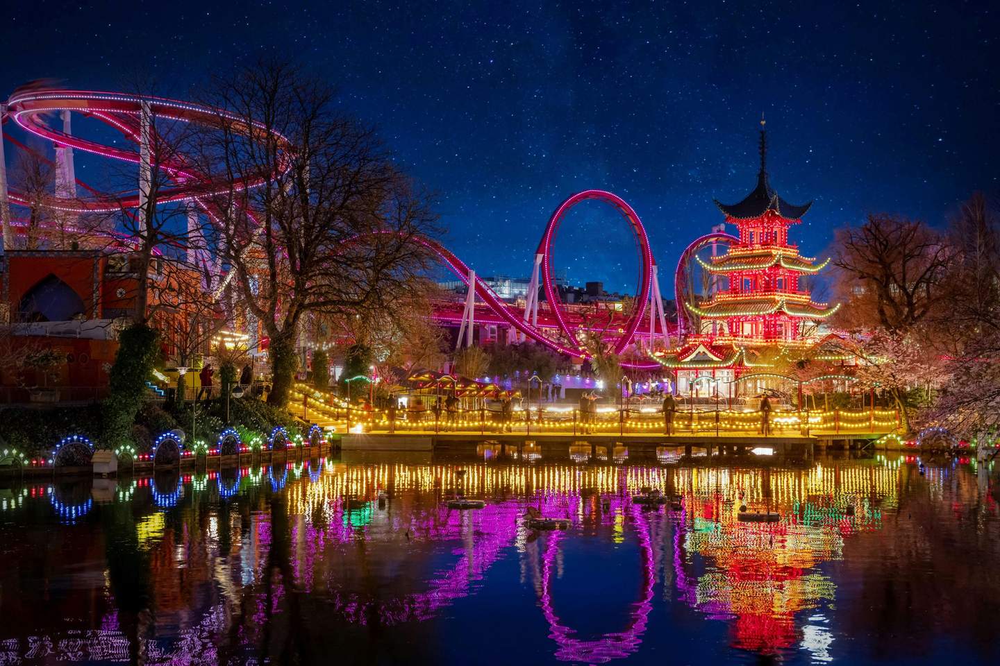 Brightly lit amusement park at night, featuring a roller coaster, pagoda, festive lights, and reflections in the water.