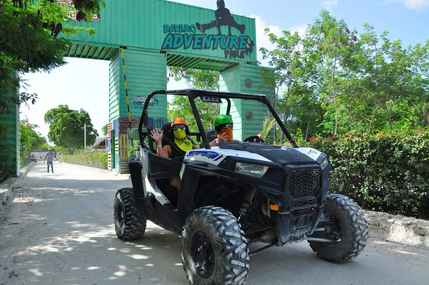 Bavaro Adventure Park Two people wearing helmets and bandanas in an off-road vehicle under an archway labeled "Bavaro Adventure Park."
