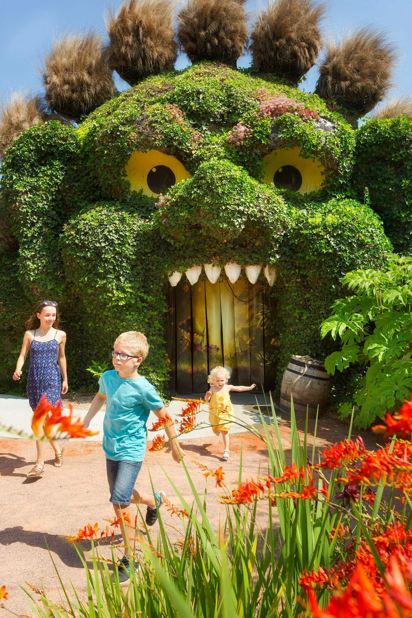 Three children and an adult walk away from a giant plant-covered sculpture with yellow eyes and a toothy mouth.