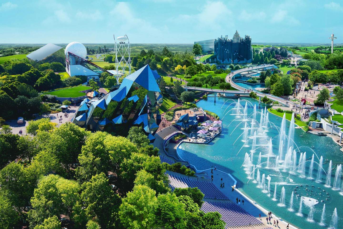 Aerial view of a theme park with futuristic buildings, water fountains, lush greenery, and visitors walking on paths.