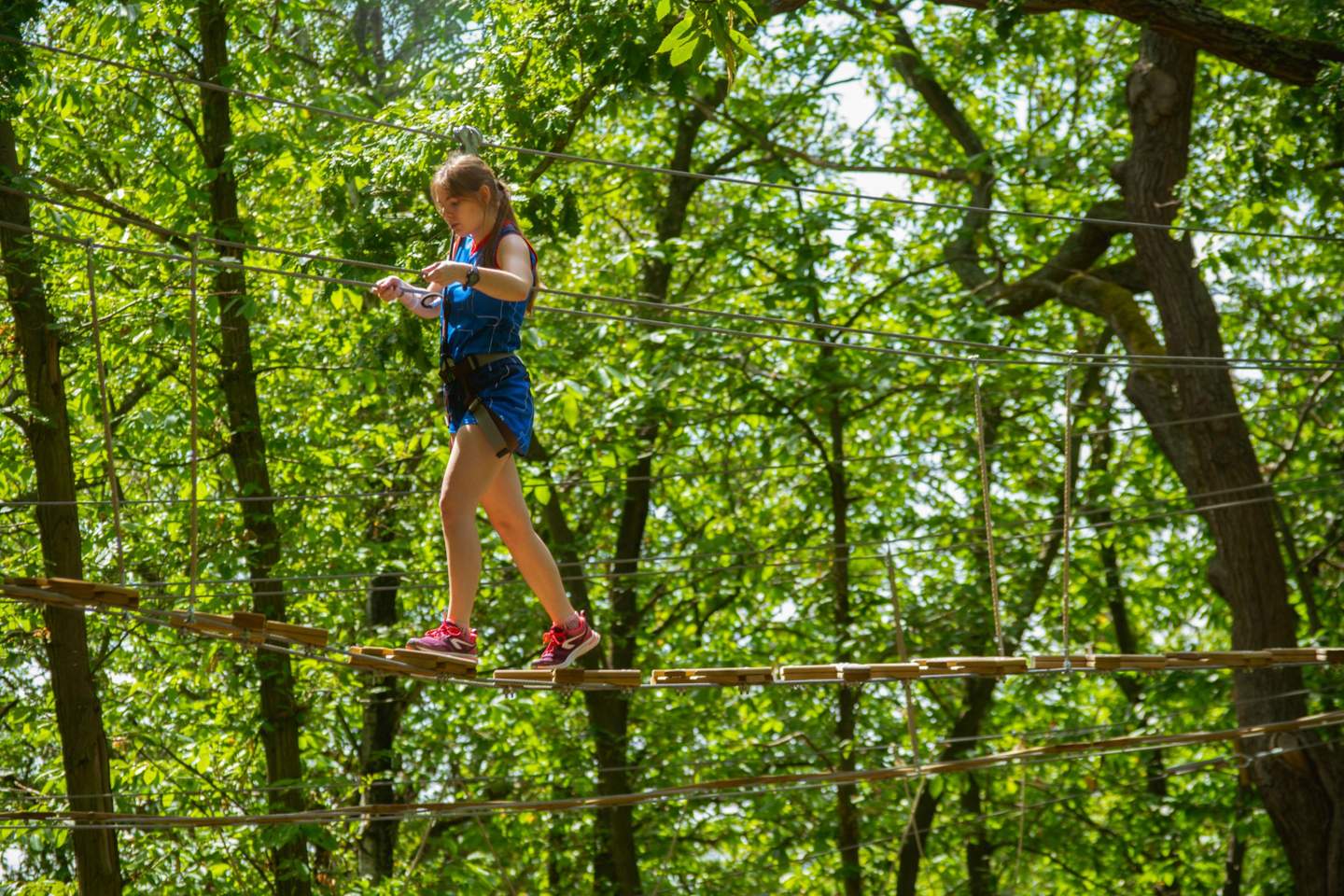 A young girl in blue gear and red shoes walks on a suspended rope bridge in a forest adventure park.