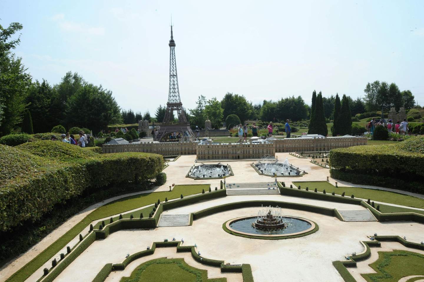 Tourists view a park with miniature replicas of the Eiffel Tower and surrounding French landmarks, with trees and bushes in the background.
